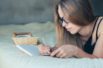 Satisfied and happy smiling girl lying on sofa in room, and writing notebook