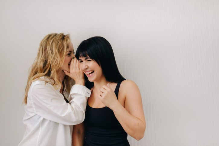 An excited woman in a black swimsuit listens to a friend in a shirt whispering in her ear on a light